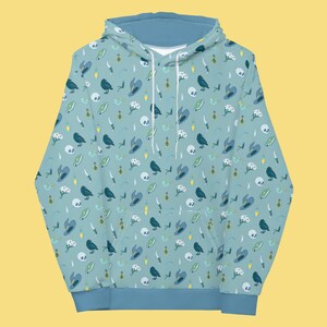 Puede incluir: Una sudadera con capucha azul claro con un patr&oacute;n repetido de cuervos negros, flores blancas, frascos de poci&oacute;n verdes, sombreros de bruja azules y dagas plateadas sobre un fondo azul claro.