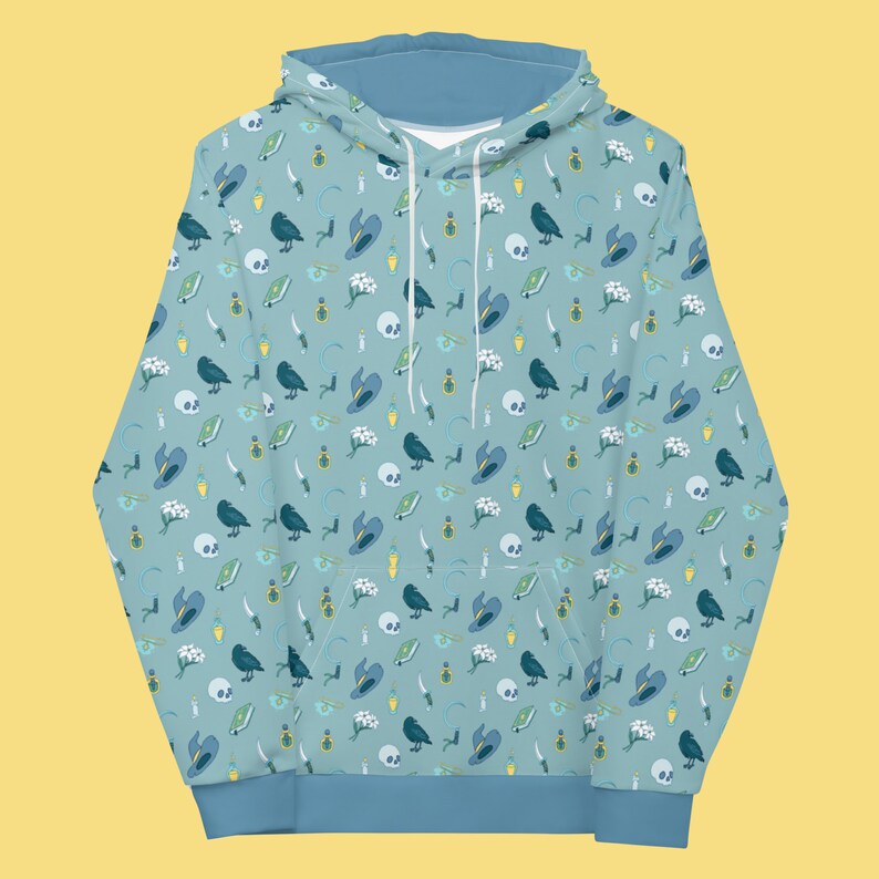 Puede incluir: Una sudadera con capucha azul claro con un patr&oacute;n repetido de cuervos negros, flores blancas, frascos de poci&oacute;n verdes, sombreros de bruja azules y dagas plateadas sobre un fondo azul claro.