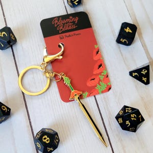 Peut inclure: Un porte-clés doré avec un pendentif en forme d'épée ornée d'une fleur de pavot rouge et de feuilles vertes. L'épée est attachée à un anneau doré et à un mousqueton. Le porte-clés est présenté sur une carte rouge avec le texte "Blooming Blades" et "Poola's Pixels".