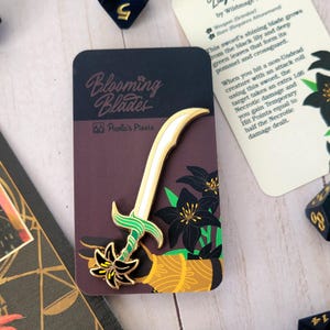 May include: A gold and green enamel pin depicting a sword with a black lily flower at the base. The pin is on a brown card with the text "Blooming Blades" and "Paola's Pixels".