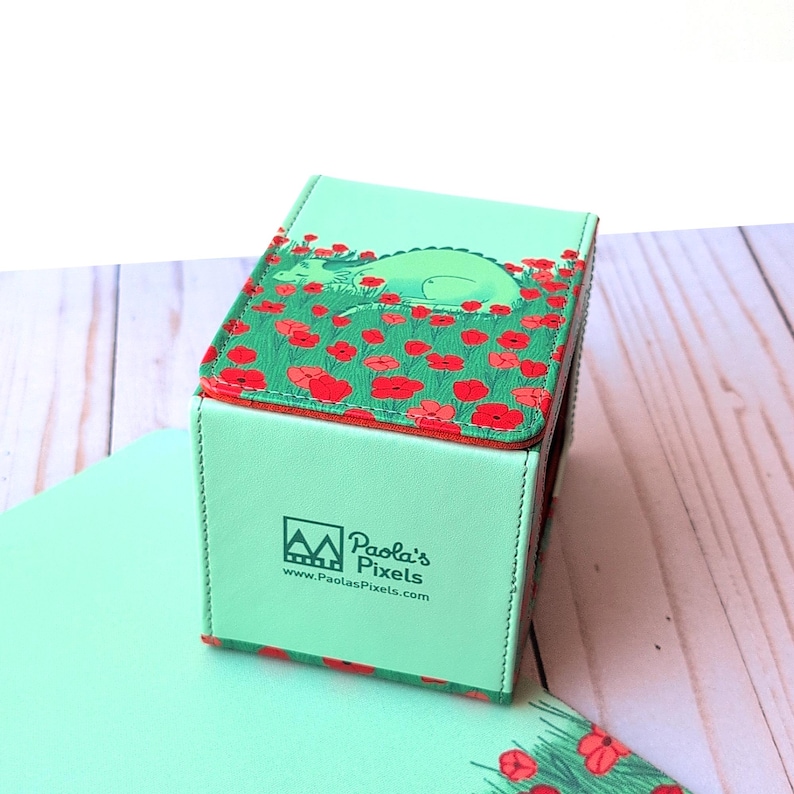 Puede incluir: Una caja verde menta con tapa abatible que presenta un drag&oacute;n durmiendo en un campo de amapolas rojas. La caja tiene el texto "Paola's Pixels" y la direcci&oacute;n web www.PaolasPixels.com.