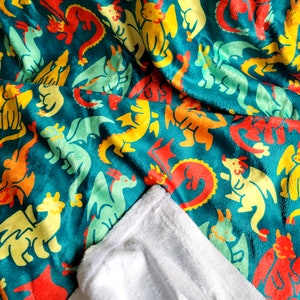 Pu&ograve; includere: Una coperta turchese con un motivo ripetuto di draghi dei cartoni animati colorati in rosso, giallo, arancione e verde. La coperta &egrave; piegata, rivelando un morbido lato inferiore bianco.