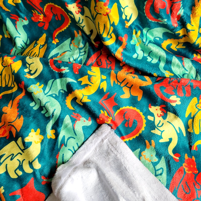 Pu&ograve; includere: Una coperta turchese con un motivo ripetuto di draghi dei cartoni animati colorati in rosso, giallo, arancione e verde. La coperta &egrave; piegata, rivelando un morbido lato inferiore bianco.