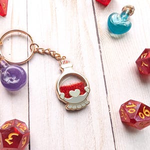 May include: Gold keychain with a red glitter potion bottle charm. The bottle has a white heart and a gold rim. The keychain is attached to a gold key ring.