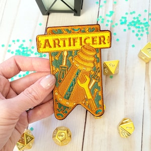 May include: Embroidered patch with the word "ARTIFICER" in gold lettering. The patch features a banner design with a gold and teal colour scheme, showcasing tools and mechanical elements. Gold dice are scattered around the patch.