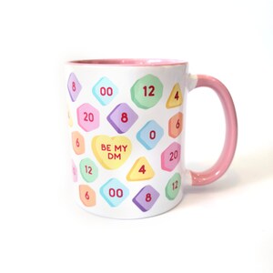 May include: White ceramic mug with a pink rim and handle. The mug features a pattern of colorful candy hearts with numbers and the text "BE MY DM" on a heart.