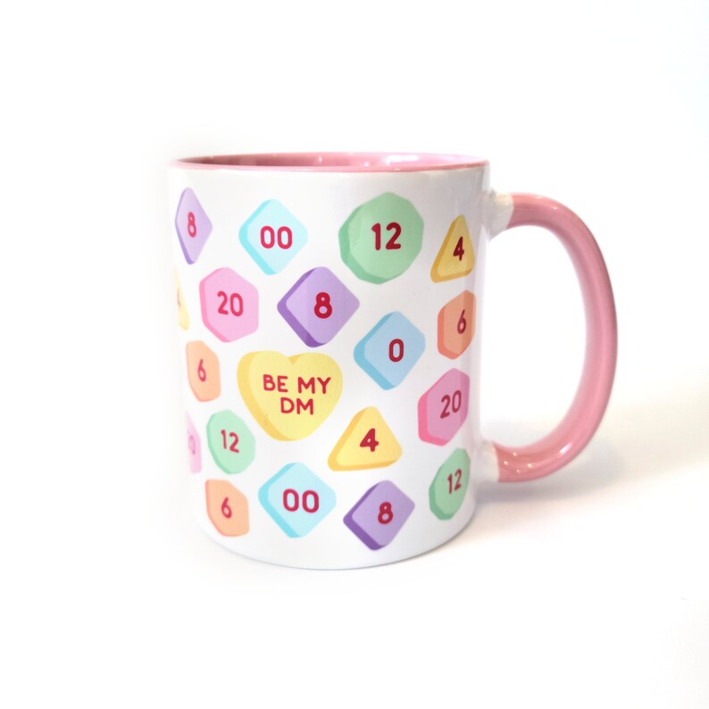 May include: White ceramic mug with a pink rim and handle. The mug features a pattern of colorful candy hearts with numbers and the text "BE MY DM" on a heart.