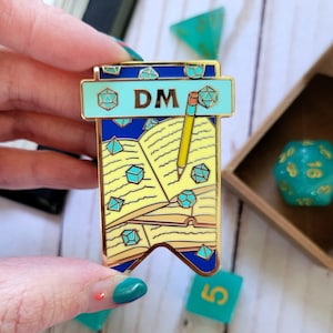 Puede incluir: Pin de esmalte dorado con un diseño azul y amarillo. El pin presenta un libro abierto con un lápiz, dados y el texto "DM".