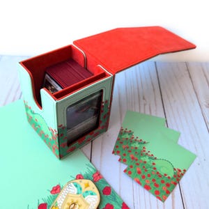 Puede incluir: Una caja de almacenamiento de cartas con interior y tapa rojos, y exterior verde claro decorado con amapolas rojas. La caja est&aacute; abierta, revelando una pila de cartas. Tambi&eacute;n hay fundas de cartas verdes a juego y un pin decorativo.