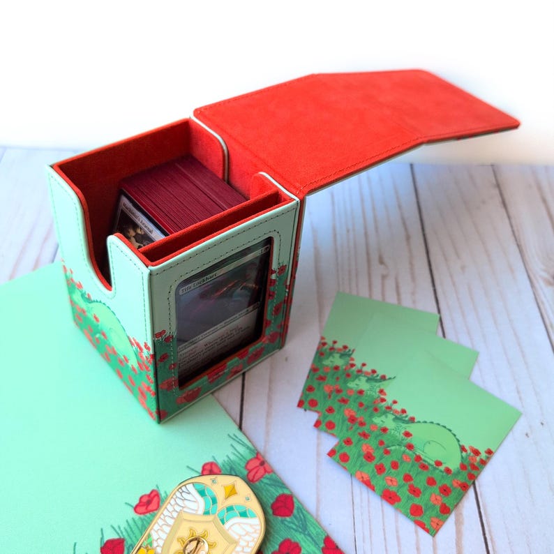 Puede incluir: Una caja de almacenamiento de cartas con interior y tapa rojos, y exterior verde claro decorado con amapolas rojas. La caja est&aacute; abierta, revelando una pila de cartas. Tambi&eacute;n hay fundas de cartas verdes a juego y un pin decorativo.