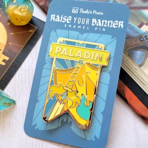 May include: A gold enamel pin with a yellow and blue banner that says "PALADIN". The banner has a sword and a flame on it. The pin is on a blue card with the text "Raise Your Banner Enamel Pin" and "Paola's Pixels".