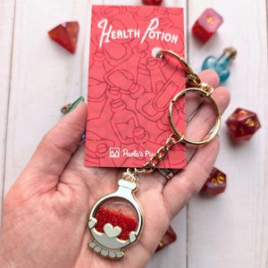 May include: Gold keychain with a red glitter filled potion bottle charm. The charm has a white heart and a gold rim. The keychain is attached to a gold chain and a gold ring. The background is a white wooden surface with a red card that says "Health Potion" and "Paola's Pixel" in white text.
