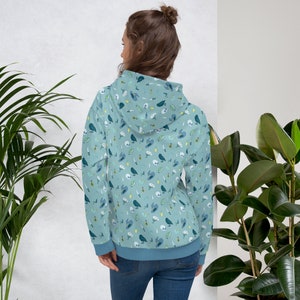 Puede incluir: Una sudadera con capucha azul claro con un patr&oacute;n de p&aacute;jaros, calaveras, flores y otros peque&ntilde;os detalles. La sudadera tiene una capucha con cord&oacute;n y un bolsillo canguro.