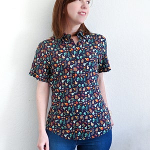 Puede incluir: Una camisa azul oscuro de botones con un estampado colorido de varios frascos de pociones, plantas y otros objetos m&aacute;gicos. La camisa tiene mangas cortas y un frente abotonado.
