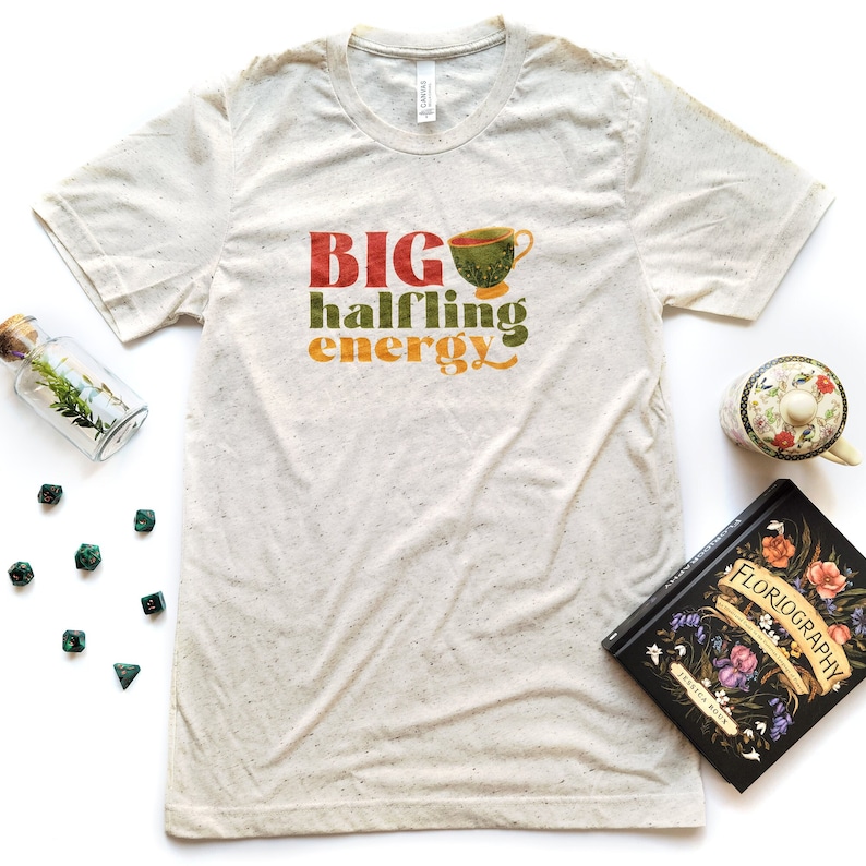 May include: A beige t-shirt with a green, orange, and red graphic that says "BIG halfling energy" with a green teacup graphic.