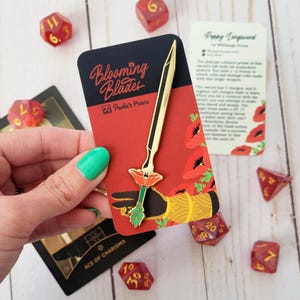 May include: A gold enamel pin depicting a sword with a red poppy flower at the hilt. The pin is on a card with the words "Blooming Blades" and "Paola's Pixels".