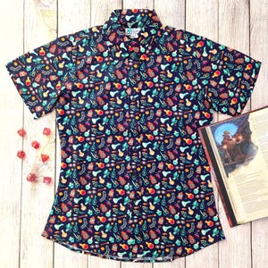Puede incluir: Una camisa azul marino de manga corta con botones y un estampado colorido de frascos de pociones, hierbas y otros objetos m&aacute;gicos. La camisa tiene un corte holgado y un dobladillo curvo.