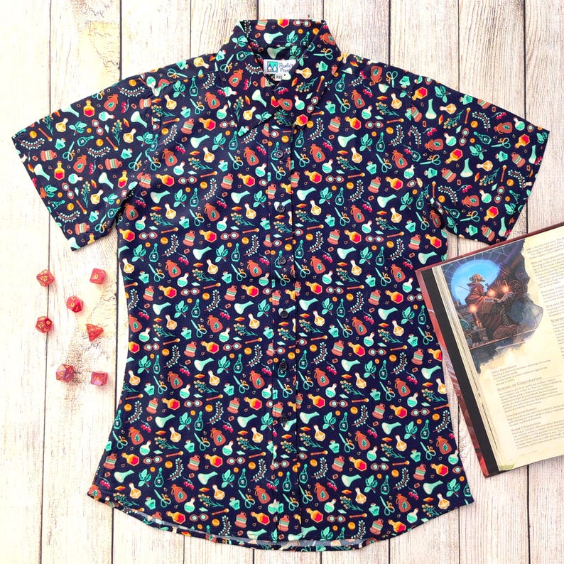 Puede incluir: Una camisa azul marino de manga corta con botones y un estampado colorido de frascos de pociones, hierbas y otros objetos m&aacute;gicos. La camisa tiene un corte holgado y un dobladillo curvo.