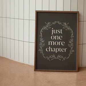May include: A framed sign with the text "just one more chapter" in white script. The text is inside a white floral border on a dark gray background. The frame is brown.