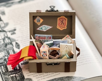 May include: A miniature suitcase filled with themed items, including a scarf, books, and treats. The suitcase is tan with stickers, and the contents are arranged on a book page. The scarf is red and gold.