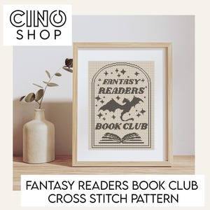 May include: A framed cross-stitch pattern featuring the text "FANTASY READERS BOOK CLUB" and a dragon silhouette. The pattern is in brown and beige tones. A white vase with a plant is to the left of the frame.