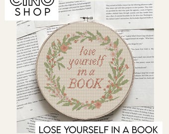 May include: A cross-stitch hoop features the phrase "lose yourself in a BOOK" in a soft, coral-colored font. The text is surrounded by a wreath of green leaves and coral-colored flowers. The design is on a beige fabric.