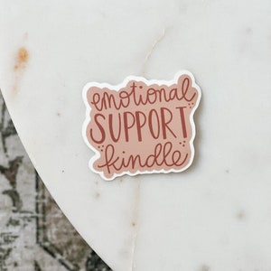 May include: A sticker with the words "emotional SUPPORT kindle" in a handwritten style. The text is in a reddish-brown color, set against a light pink background. The sticker has a white border and is placed on a white marble surface.
