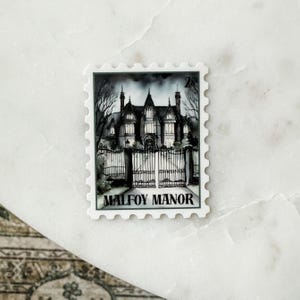 May include: A black and white stamp featuring Malfoy Manor. The image shows a detailed illustration of a gothic mansion behind a wrought iron gate. The stamp has a white border and the text "MALFOY MANOR" is printed at the bottom.