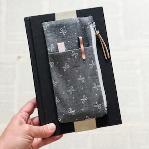 May include: A gray fabric pencil case with a white bow pattern and a zipper, attached to a black book. The pencil case has a pocket holding a pink eraser and a copper-colored pen. The book has a beige bookmark.
