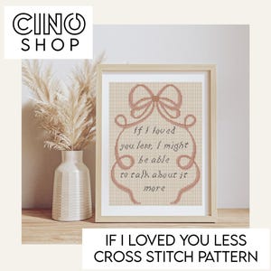 Op de afbeelding: Een ingelijst kruissteekpatroon met de tekst "If I loved you less, I might be able to talk about it more." Het ontwerp heeft een strik en een decoratieve rand in een zachte roze tint. Het frame is licht van kleur.