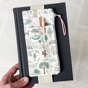 May include: A black book with a patterned fabric bookmark and pencil case. The fabric features a garden scene with trees, a pond, and a gazebo. A pink pen and highlighter are in the pencil case. The bookmark has a pink ribbon.