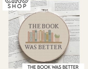 May include: A cross-stitch pattern on a beige fabric hoop. The design features the text "THE BOOK WAS BETTER" above a row of stylized books and a small potted plant. The pattern is suitable for embroidery.