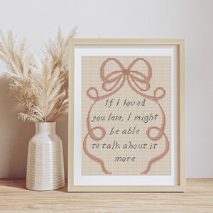 Op de afbeelding: Ingelijst kruissteekkunstwerk met een lichtbeige achtergrond. Het ontwerp bevat een strik en de tekst "If I loved you less, I might be able to talk about it more". Links van het frame staat een vaas met gedroogde bloemen.