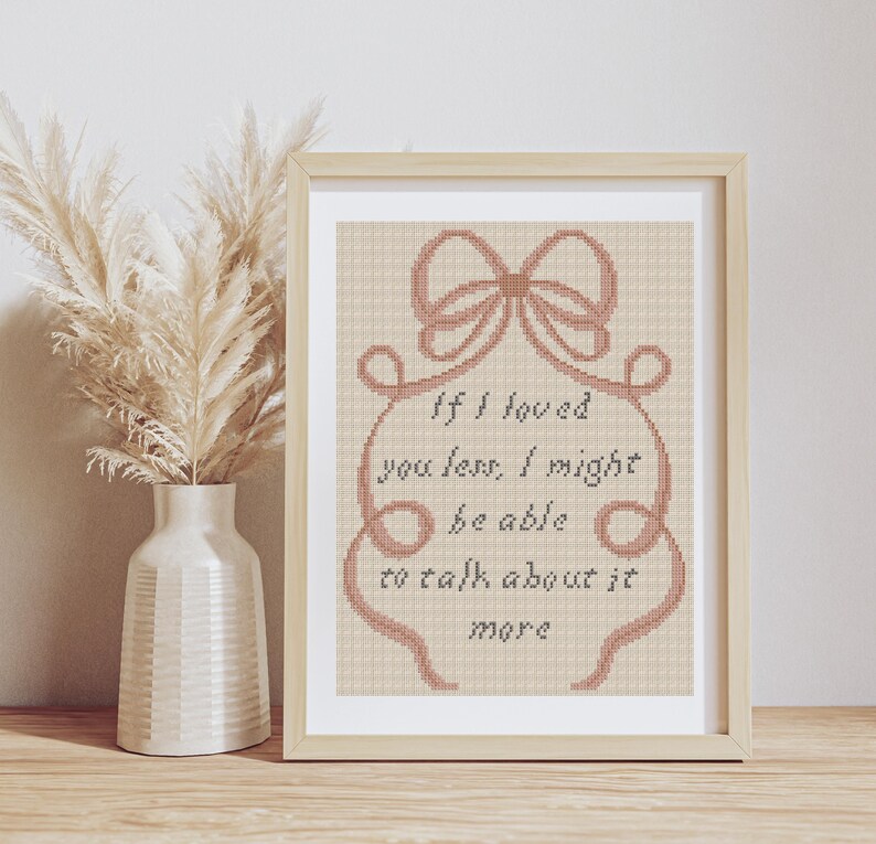 Op de afbeelding: Ingelijst kruissteekkunstwerk met een lichtbeige achtergrond. Het ontwerp bevat een strik en de tekst "If I loved you less, I might be able to talk about it more". Links van het frame staat een vaas met gedroogde bloemen.