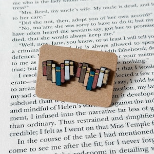 May include: A pair of wooden stud earrings shaped like stacks of books. Each book has a different color spine, including brown, teal, and white. The earrings are on a small, brown card, with a book page in the background.