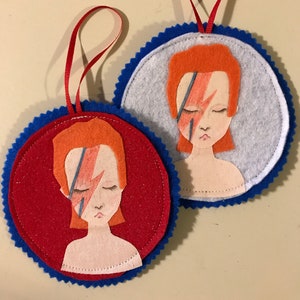 Puede incluir: Dos adornos de fieltro con ribete azul, cada uno con un retrato de fieltro rojo y naranja de David Bowie con un rayo en la cara. Los adornos tienen cintas rojas y azules para colgar.