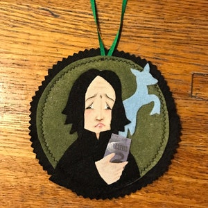 Op de afbeelding: Een vilten ornament met een cartoon afbeelding van Severus Snape die een Patronus-hanger vasthoudt. De hanger is een blauw hert. Het ornament is groen en zwart met een zwarte gegolfde rand.