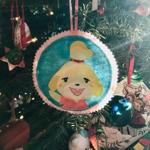 May include: A round Christmas tree ornament featuring a cartoon character with a yellow head and a pink shirt. The ornament has a light blue center, a pink scalloped edge, and a pink ribbon for hanging. The ornament is hanging on a green Christmas tree.