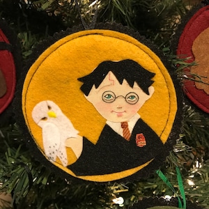 Puede incluir: Un adorno navideño de fieltro con Harry Potter y su búho Hedwig. Harry lleva su uniforme de Hogwarts con el escudo de Gryffindor. El adorno es redondo y tiene un fondo amarillo.