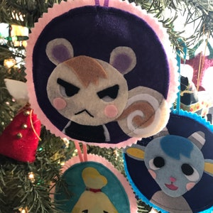 May include: Handmade felt Christmas ornaments featuring characters from a popular video game. Each ornament is round, with a character's face on a dark background, framed by a pink scalloped edge. Purple ribbon loops for hanging.