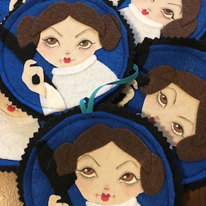 May include: Four blue felt ornaments with a white felt portrait of a woman with brown hair and a white shirt. The woman is holding a black felt object in her hand.