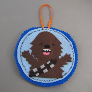 May include: A blue and brown felt ornament featuring a cartoon Chewbacca character with a big smile. The ornament has a loop of orange felt for hanging.