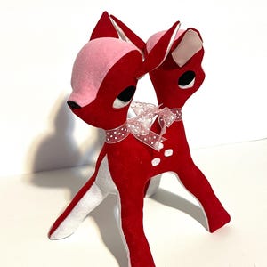 May include: A pair of red deer figurines with pink and white accents. The deer have black eyes and are adorned with a sheer, polka-dot ribbon around their necks. The figurines are made of a soft, plush material and are standing on a white surface.