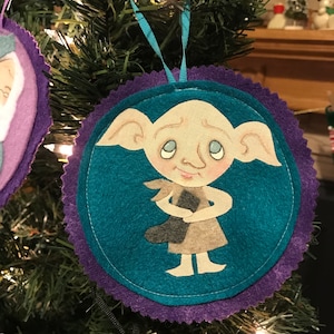 May include: A purple and teal felt Christmas ornament featuring a hand-painted illustration of Dobby the house-elf from the Harry Potter series. Dobby is wearing a grey and brown outfit and has a big smile on his face.