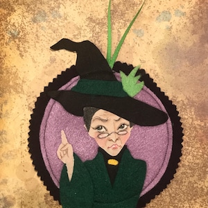 May include: Felt ornament of a witch wearing a black hat and green robes, with a purple background and a black border.