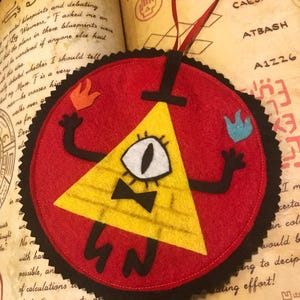 Puede incluir: Adorno de fieltro rojo con un triángulo amarillo, un solo ojo, brazos negros y una pajarita. Tiene un borde negro festoneado y está colocado sobre un libro abierto con texto manuscrito y la palabra "CODE". Se adjunta una cinta roja para colgarlo.