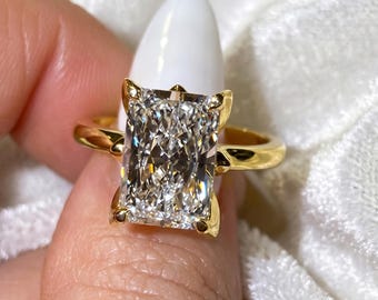 May include: A radiant-cut diamond ring with a gold band. The large, rectangular diamond is set in a gold prong setting. The ring is displayed against a soft, white background, highlighting the brilliance of the gemstone.