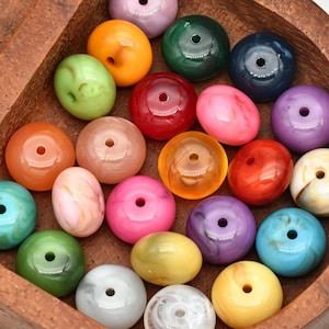 Puede incluir: Una colección de cuentas redondas en forma de disco en una variedad de colores sólidos y jaspeados, incluyendo rojo, azul, verde, amarillo, rosa, naranja y morado. Cada cuenta presenta un orificio central y están dispuestas en un recipiente de madera.