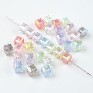 Pode incluir: Uma coleção de pequenas contas cúbicas translúcidas com um acabamento iridescente. As contas apresentam uma superfície facetada que reflete a luz em várias cores pastel, incluindo rosa, azul, amarelo e verde. Uma agulha prateada enfia várias contas.