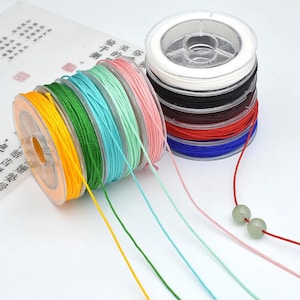 May include: A collection of nylon cord spools in various colors including yellow, green, teal, light green, pink, white, black, brown, red, and blue. Two light green beads are threaded onto the red cord extending from the blue spool.